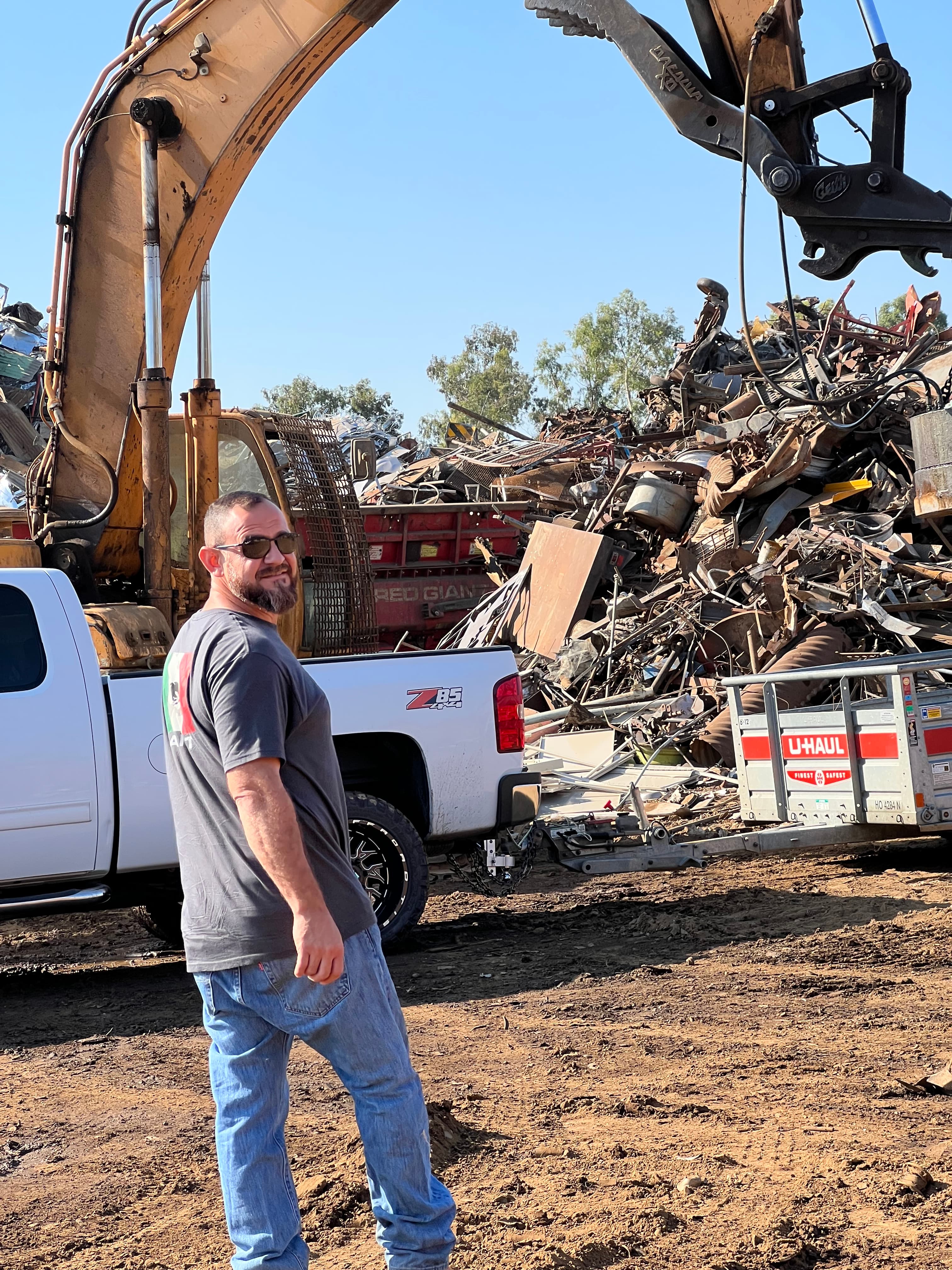 Man in a scrapyard with a large scrap metal pile, excavator arm, and pickup truck.