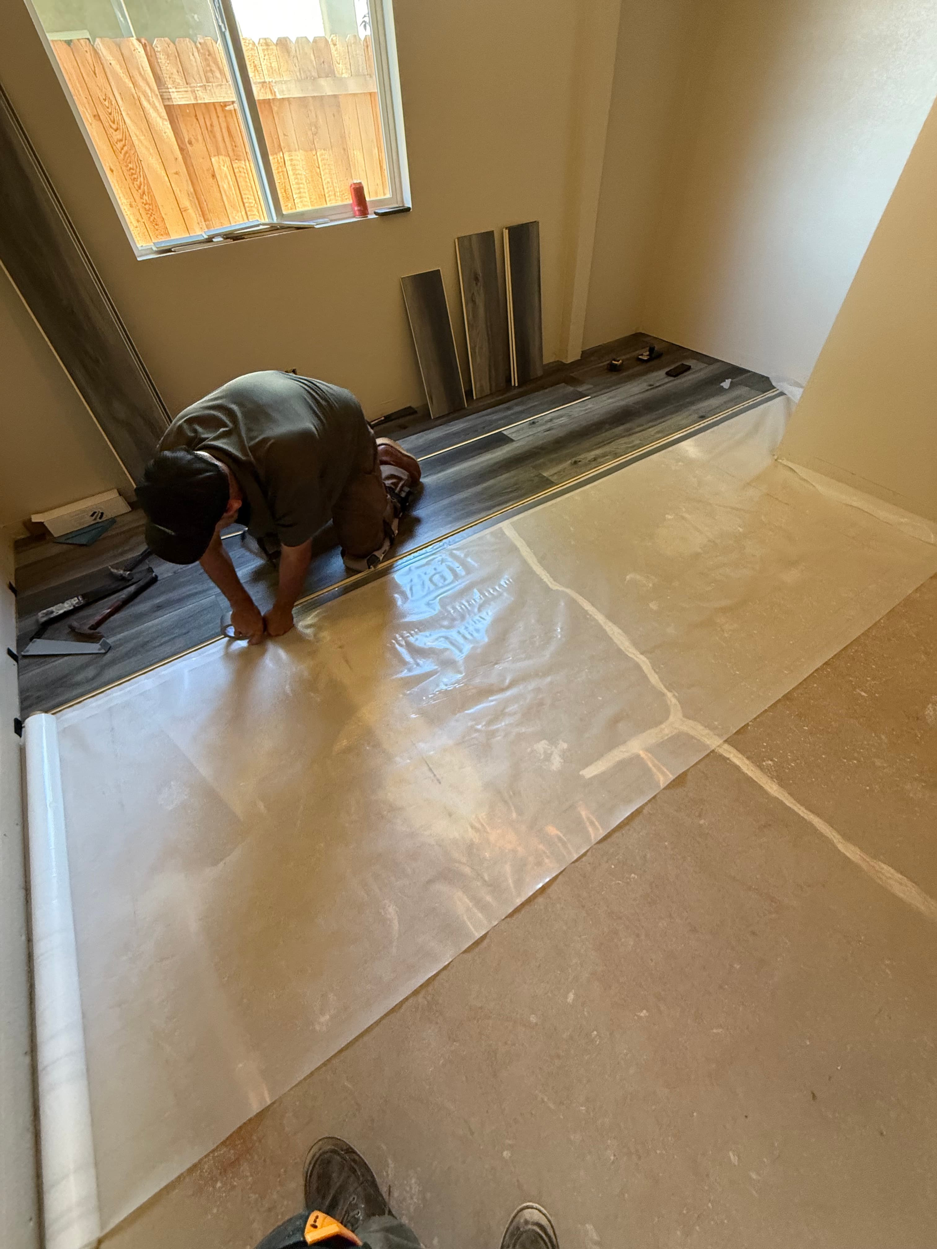 A man installs dark wood-look laminate flooring over a plastic moisture barrier on a subfloor.